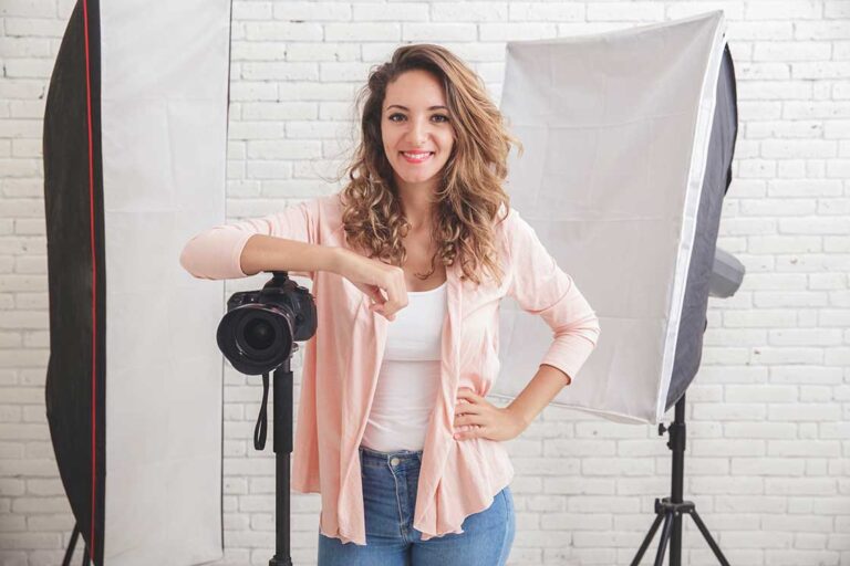 Femme souriante en studio, posant avec son appareil photo sur un trépied, entourée d’éclairages professionnels. Illustration de la transition vers une carrière de photographe de mariage.