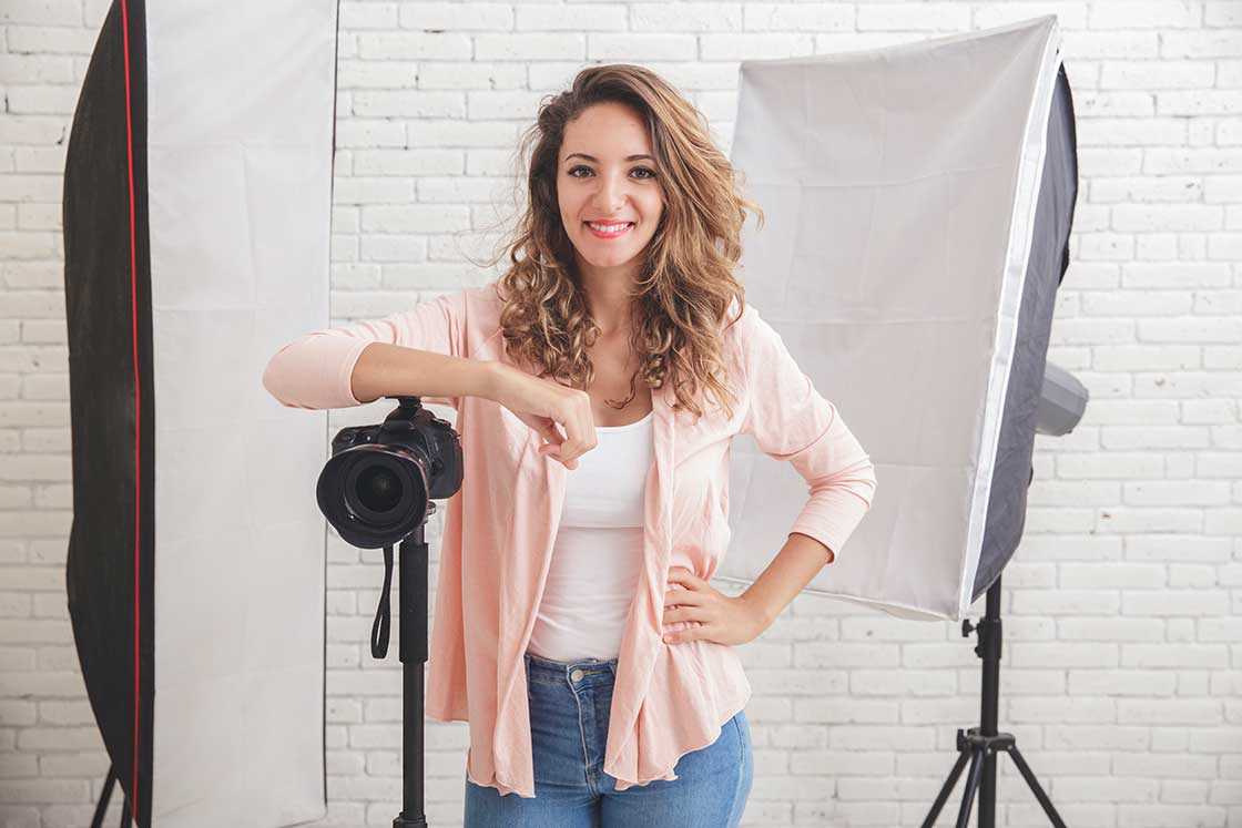 Femme souriante en studio, posant avec son appareil photo sur un trépied, entourée d’éclairages professionnels. Illustration de la transition vers une carrière de photographe de mariage.