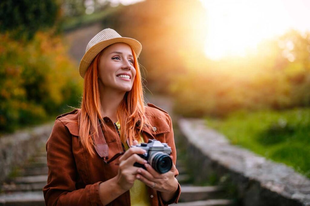 Femme souriante tenant un appareil photo, vêtue d’une veste marron et d’un chapeau, profitant d’une belle lumière en extérieur. Illustration de la liberté et du plaisir de devenir photographe de mariage.